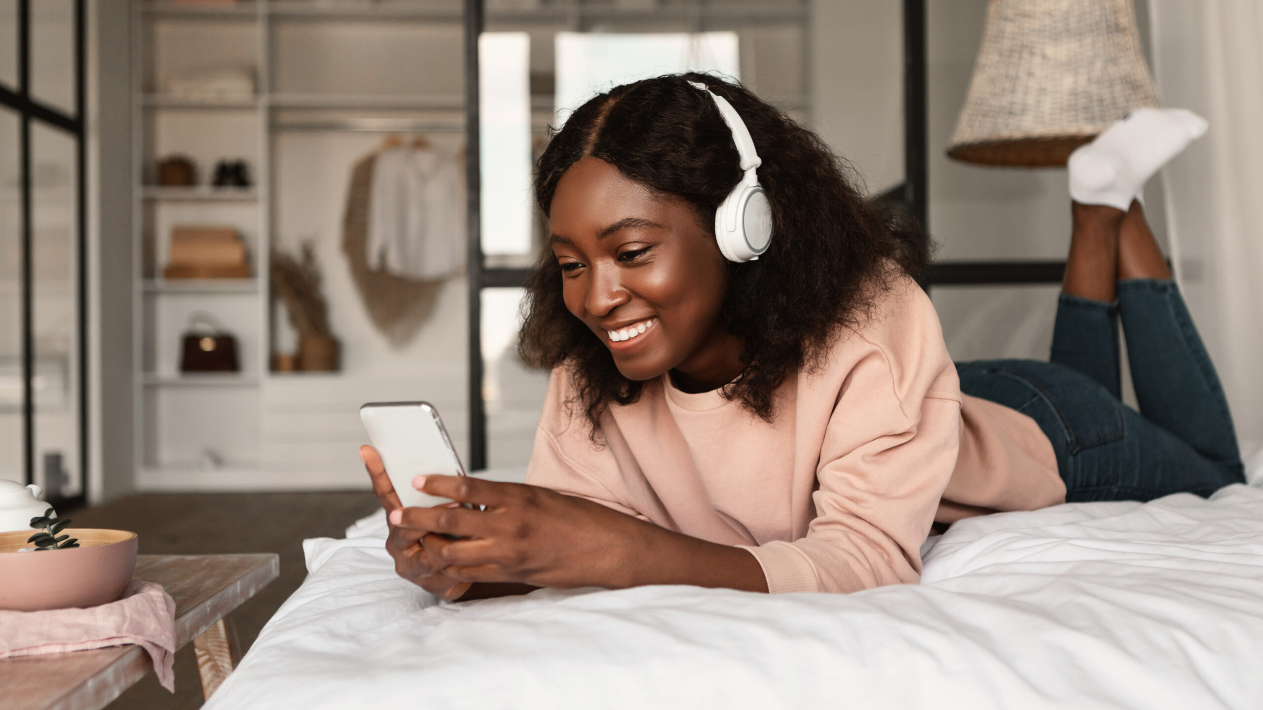 Happy African Lady Using Phone Wearing Headphones Lying In Bedroom